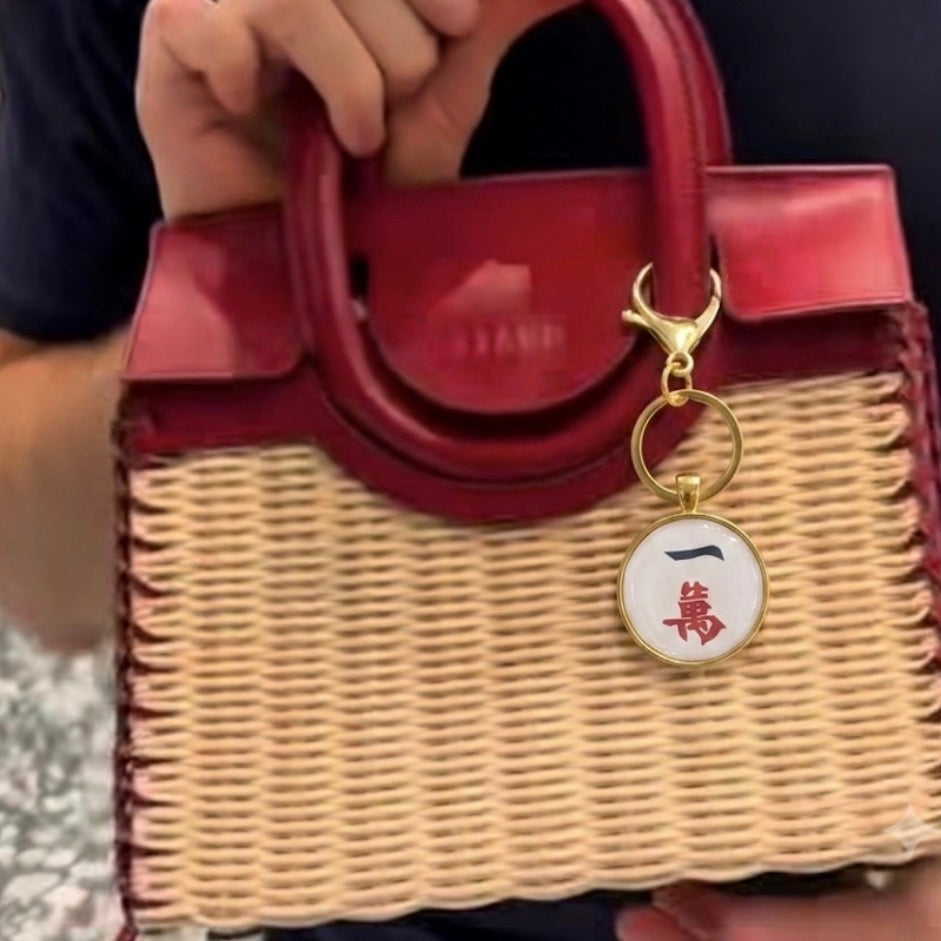 A structured handbag with a glossy dark red leather top and handles, combined with a natural woven straw lower body. It features a gold-tone keyring charm with a white circular tag printed with a red Chinese character.